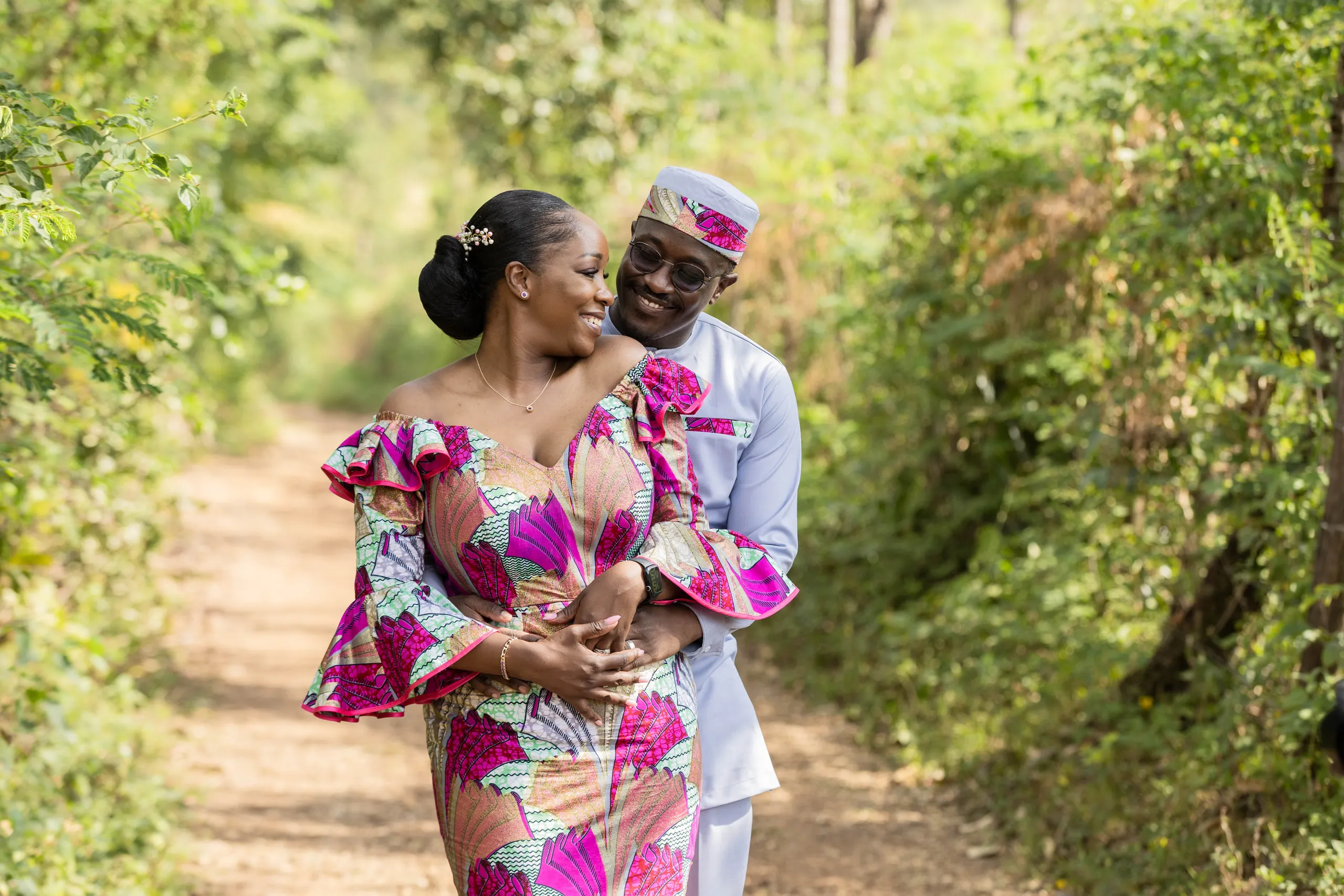Couple on a forest path, she in a pink print dress, he in white with a matching cap