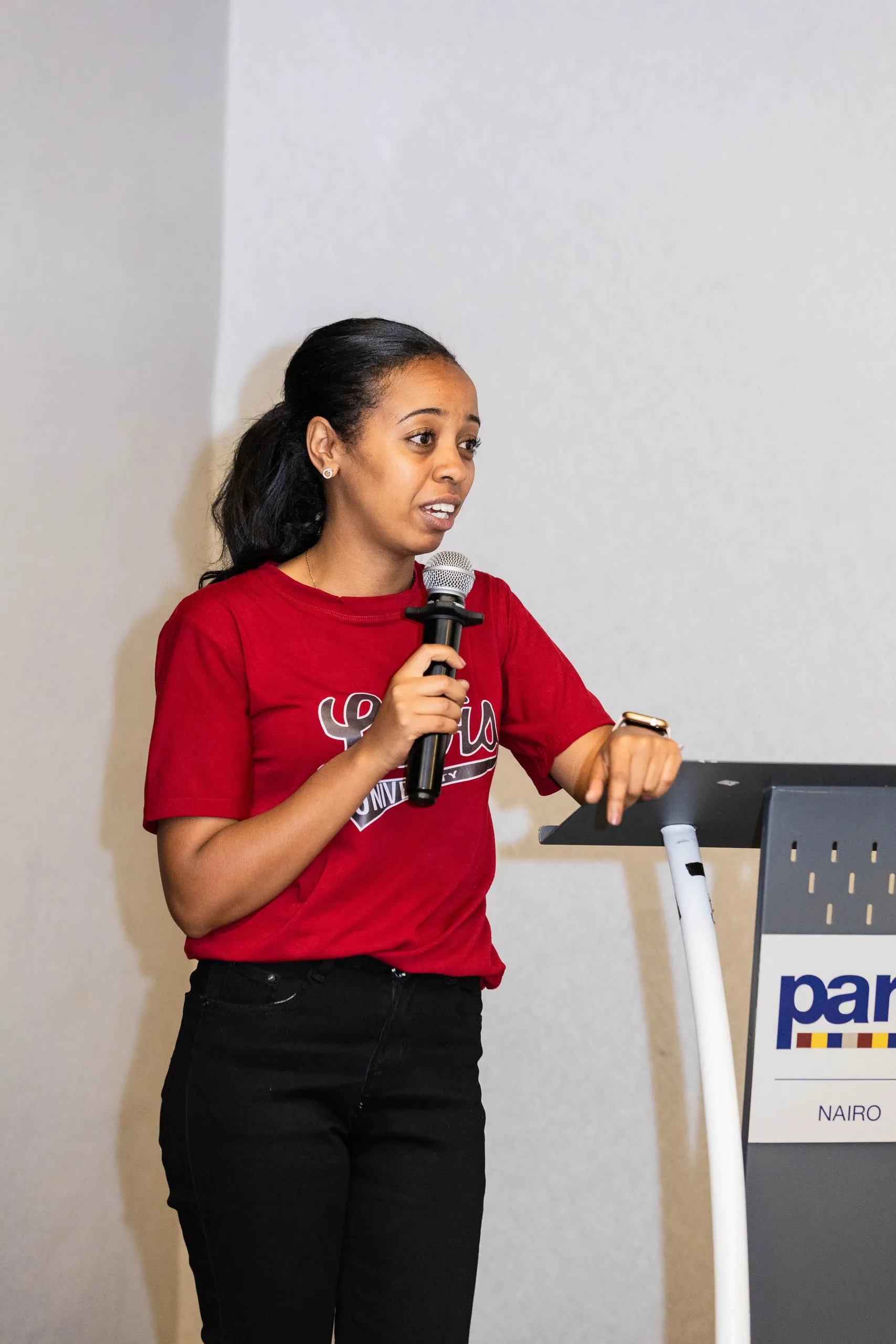 Woman in a red university tee holding a microphone and speaking next to a lectern