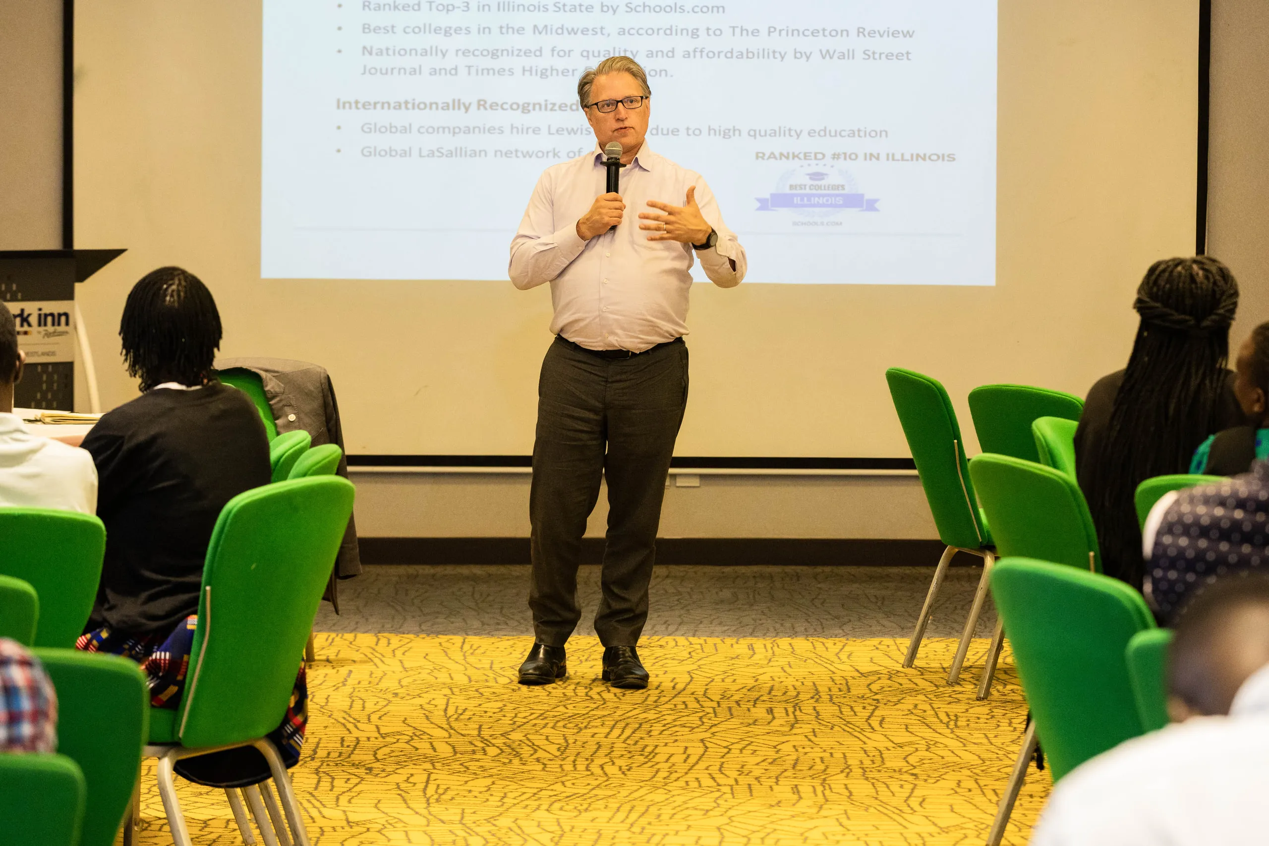 Man in a pale shirt addressing rows of green chairs beside a projected slide