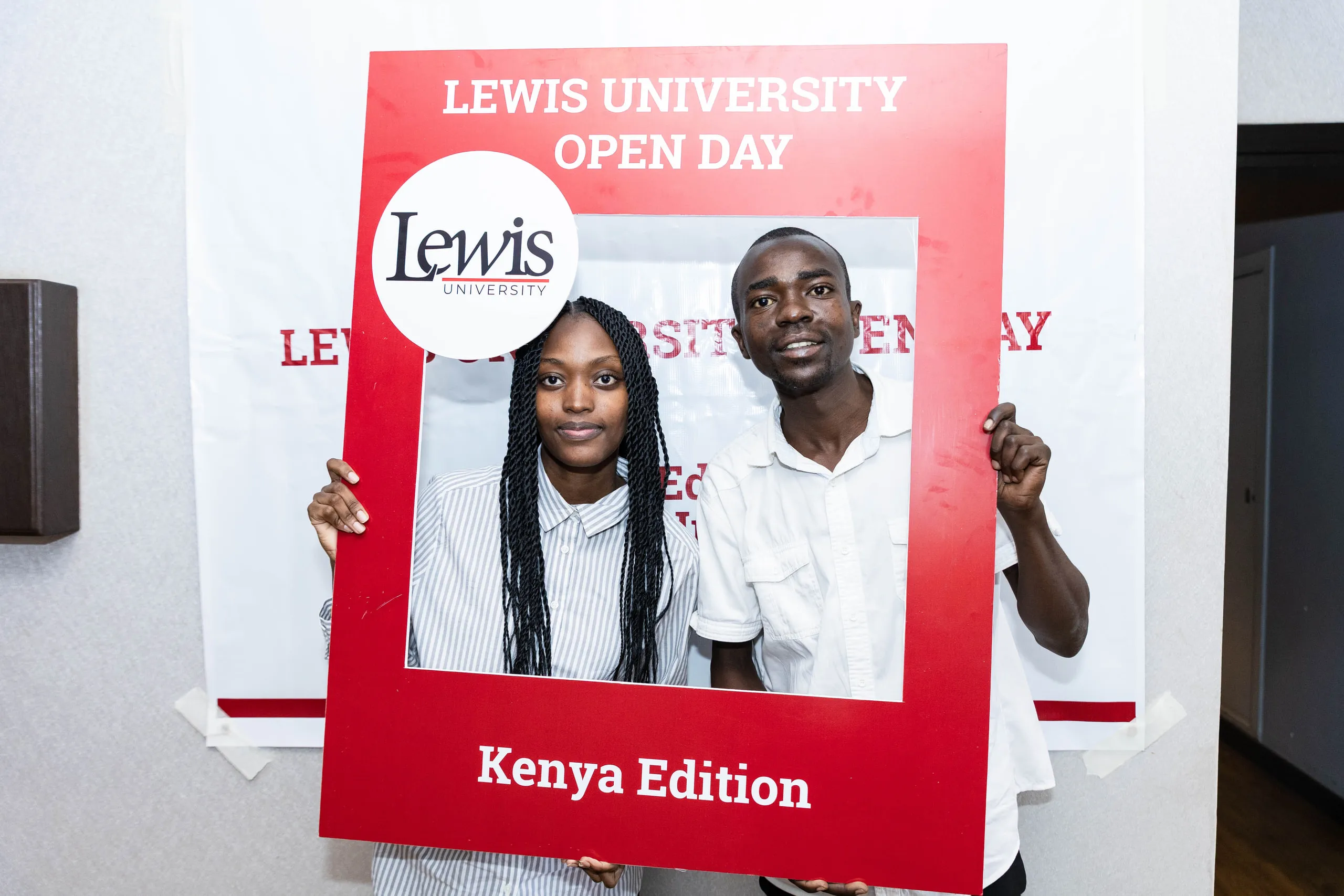 Two young people holding a red open day photo frame in front of a banner