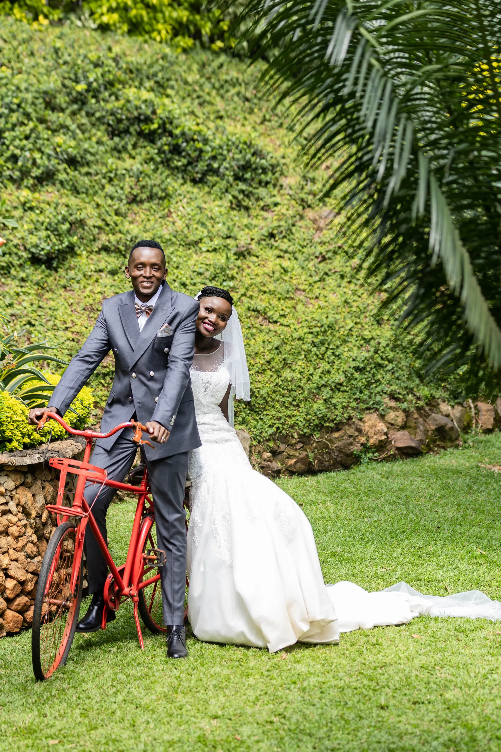 Bride and groom with a red bicycle on a lawn below a green hillside