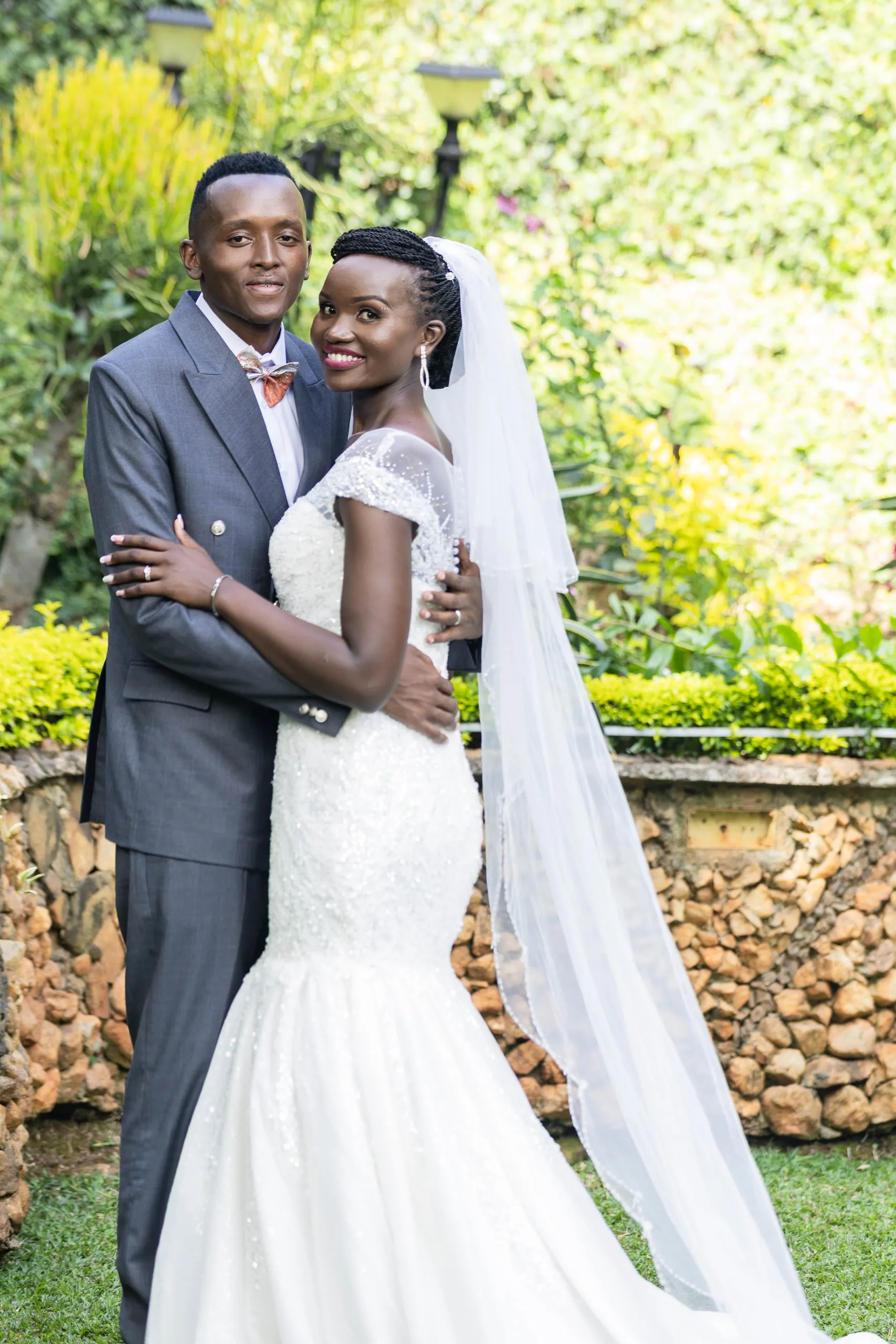 Bride and groom embracing in front of a stone wall, her veil to the ground