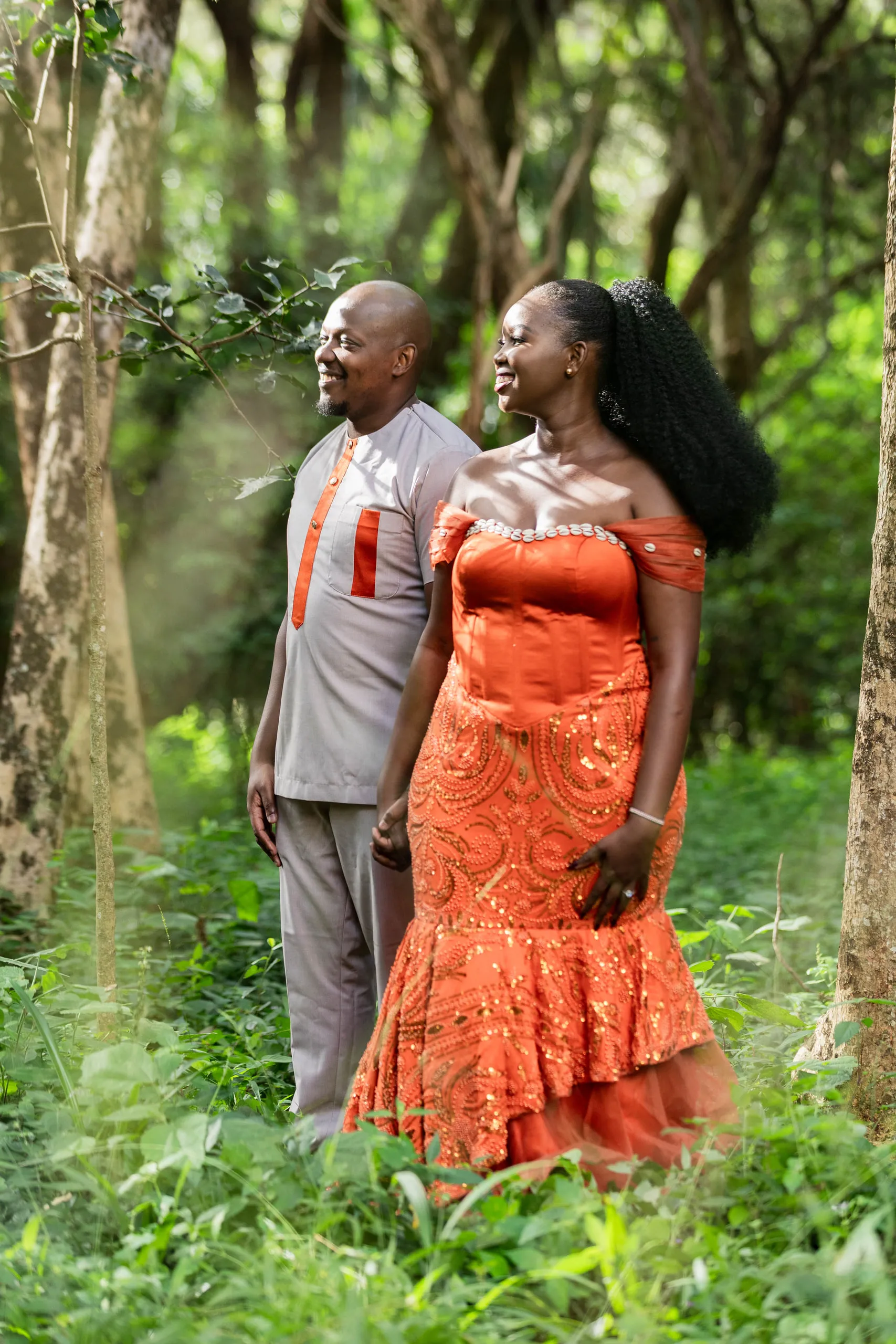 Couple among tall trees, she in an orange lace gown, he in grey with orange trim