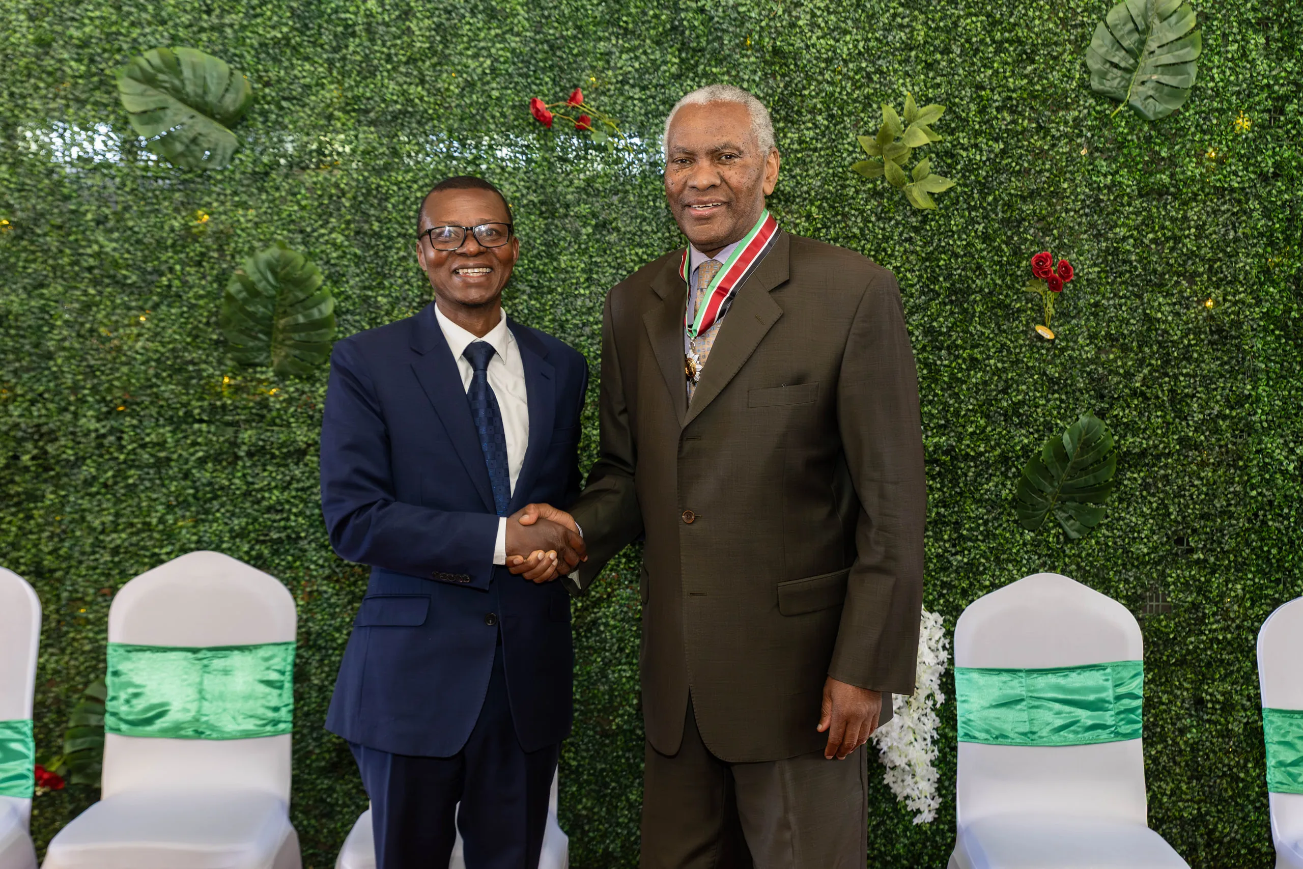 Two men shaking hands in front of a green leaf wall, one wearing a ribbon with a medal