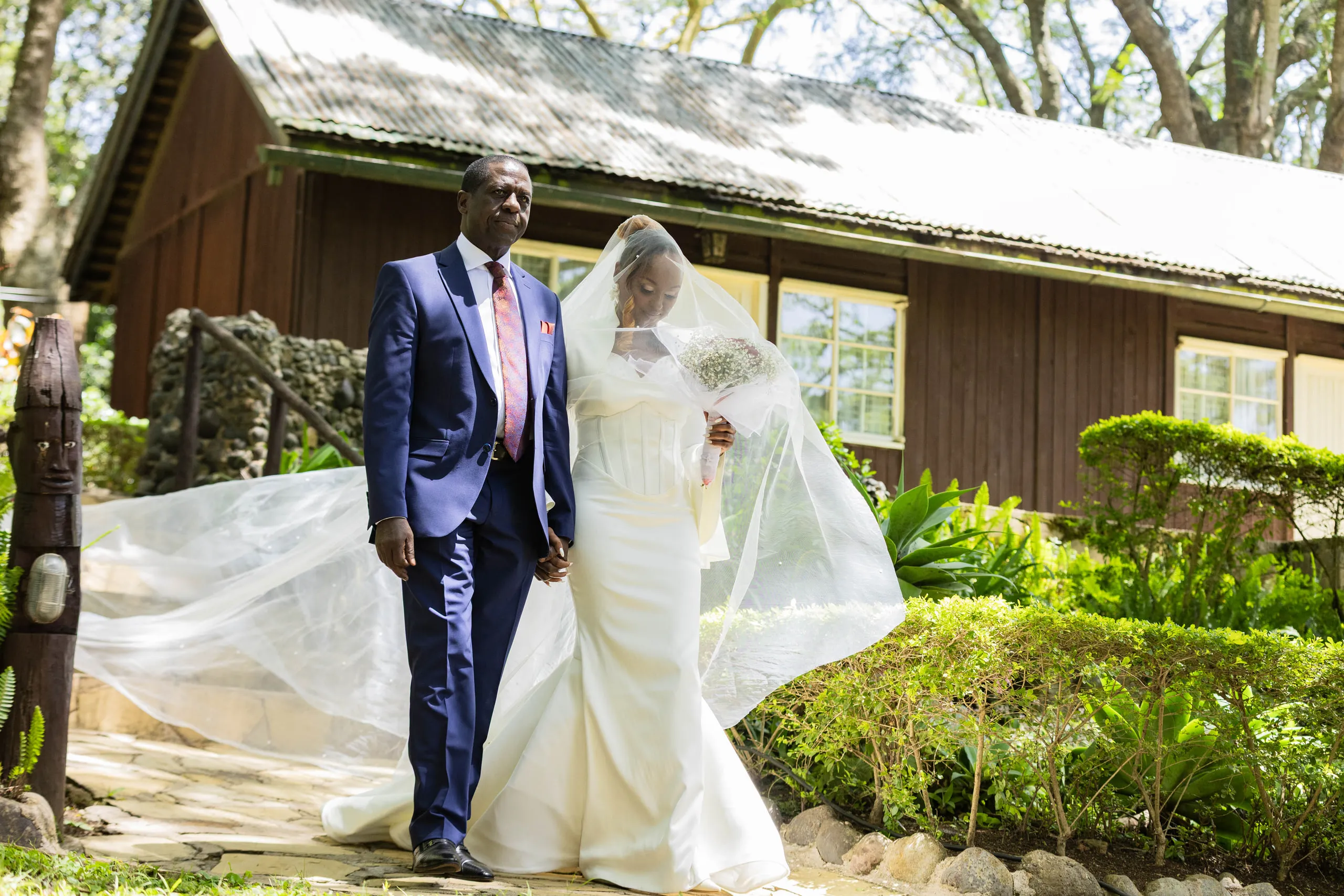 Bride walking arm in arm with a man in a navy suit, her veil trailing