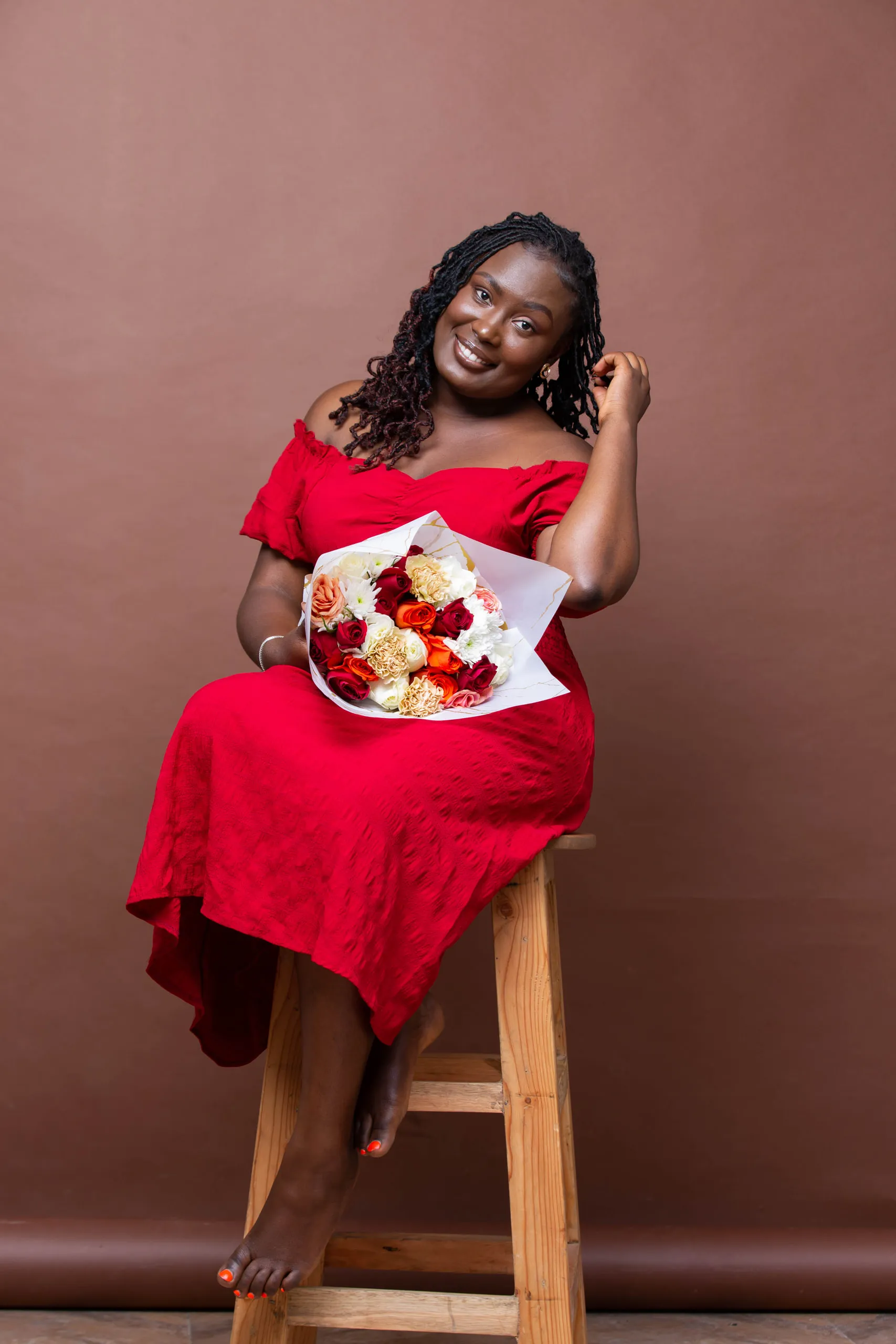 Woman in a red dress on a wooden stool holding a bouquet, against a brown backdrop