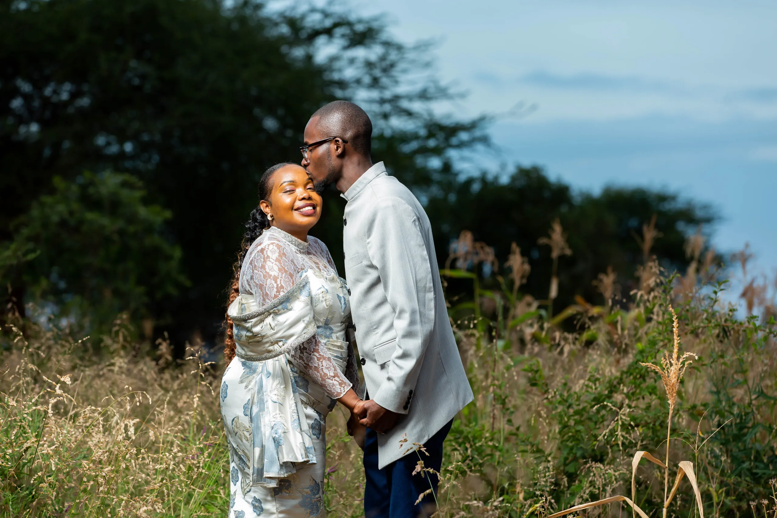 Groom kissing the bride's forehead in tall grass, she in silver and blue lace