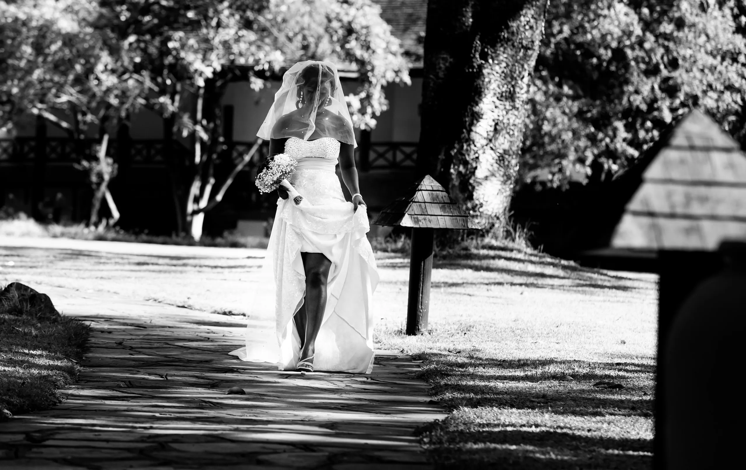Bride lifting her dress as she walks down a garden path, in black and white