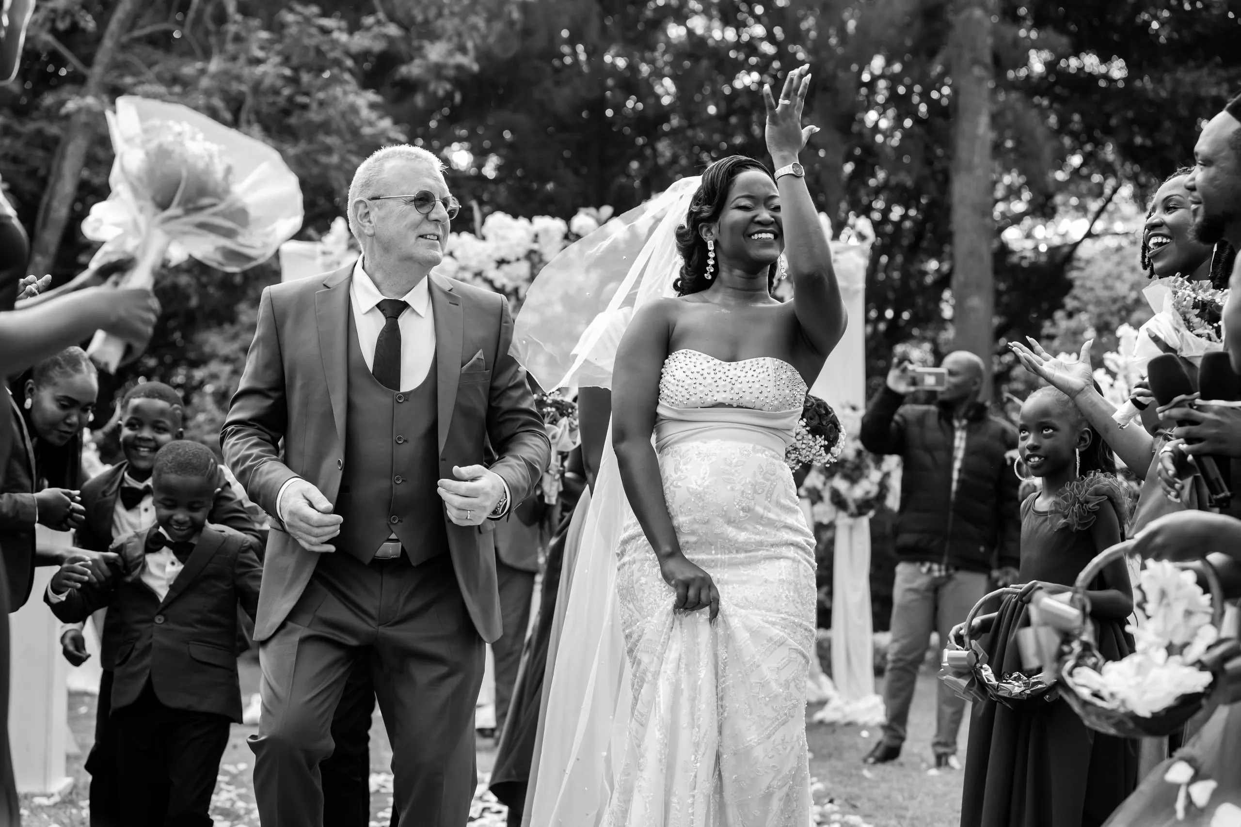 Bride raising her hand as guests throw petals, in black and white