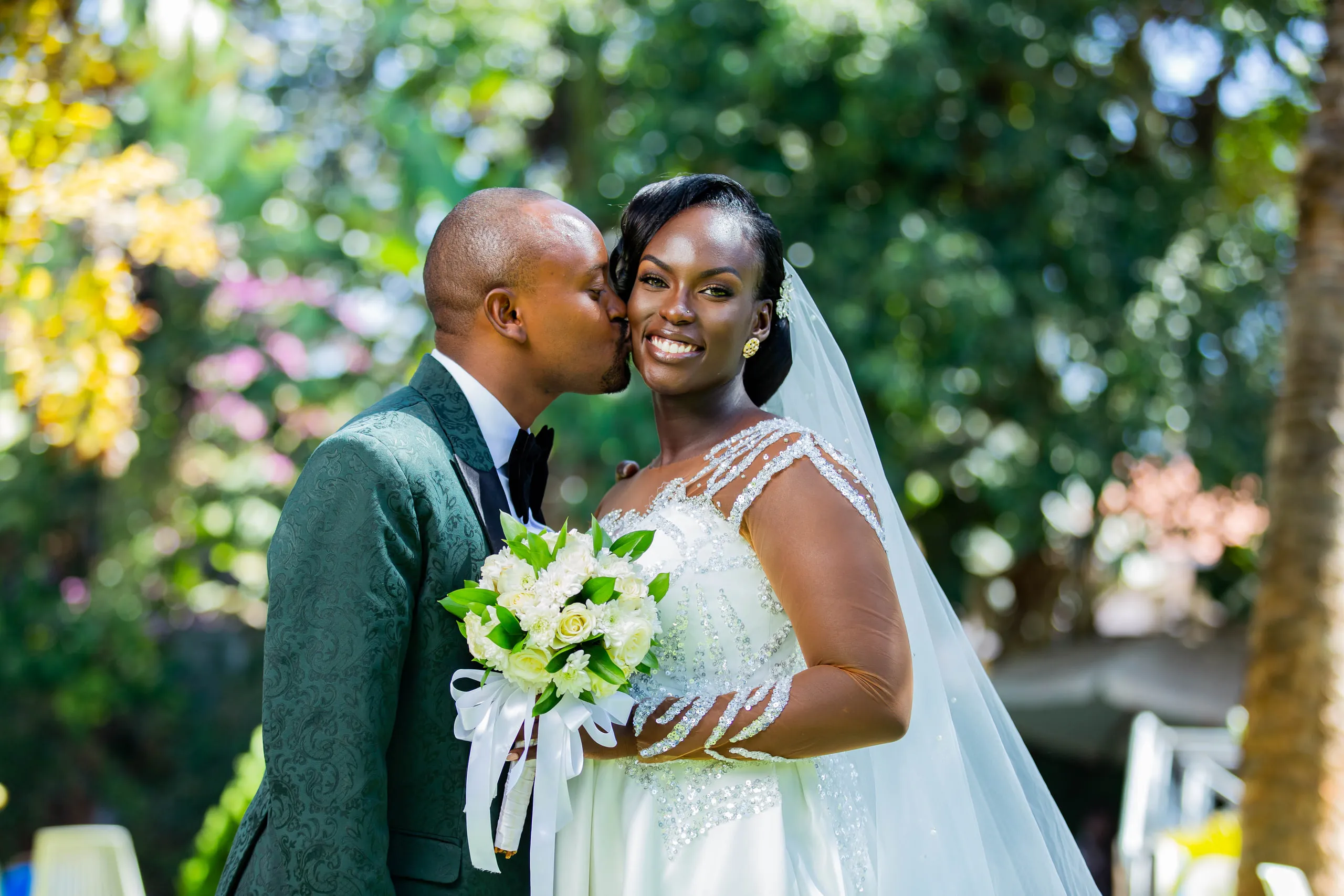 Groom in a green jacket kissing the bride's cheek as she holds white roses