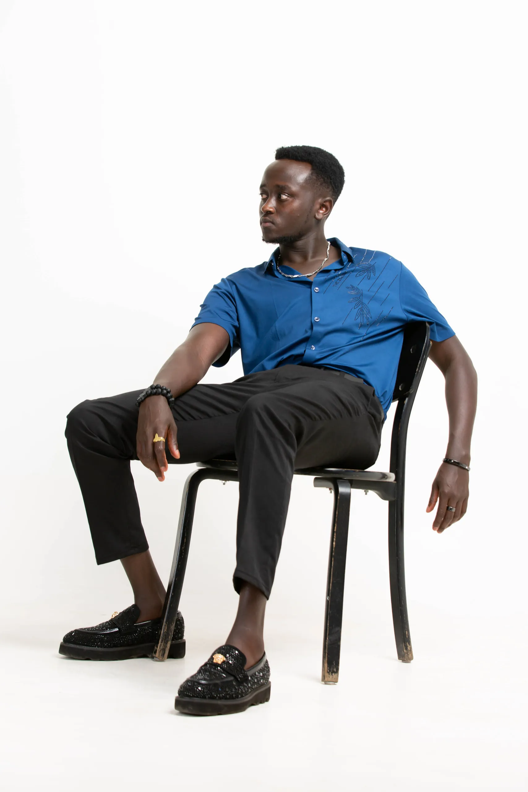 Man in a blue shirt and black loafers sitting back on a chair, looking to one side