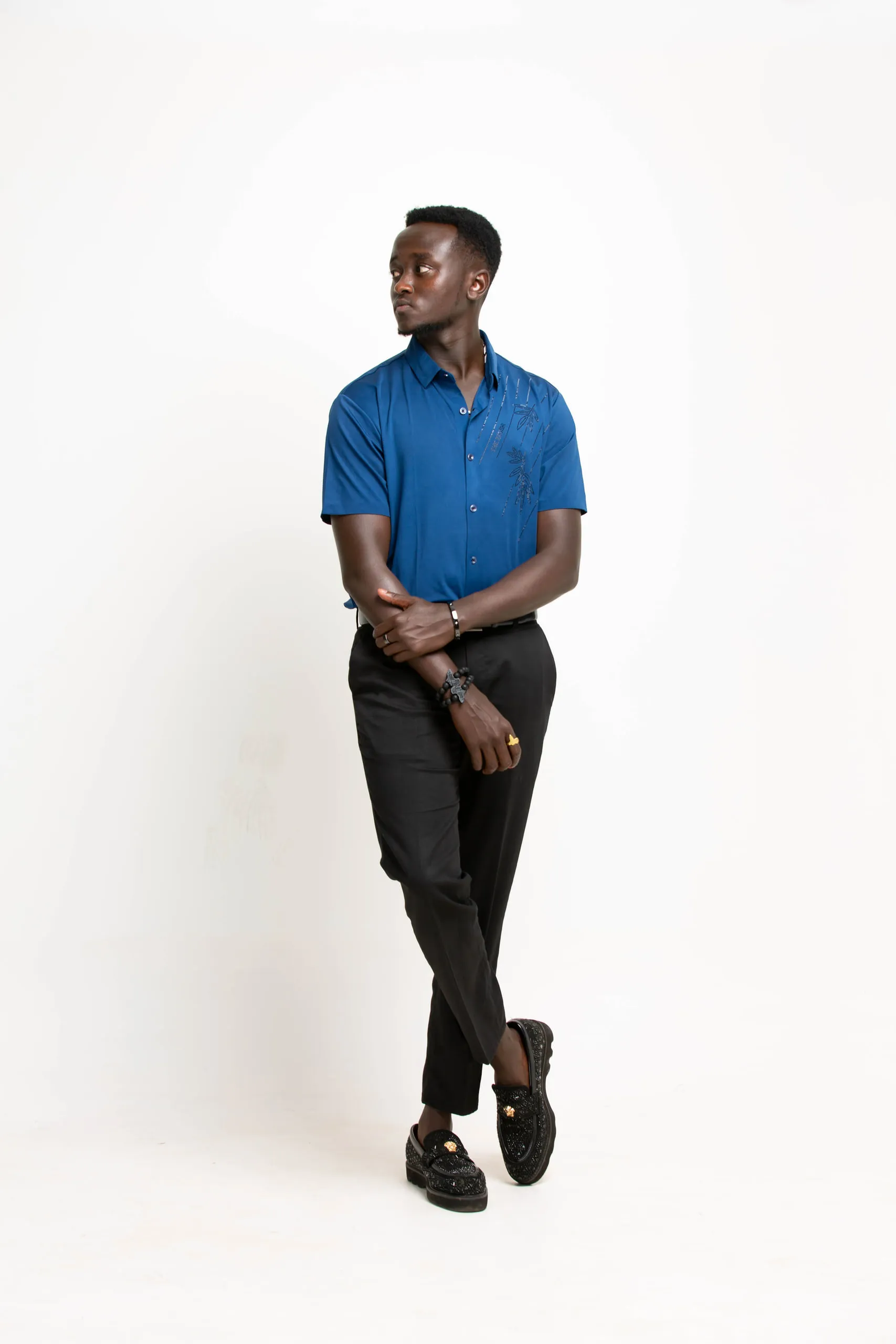 Man in a blue shirt and black trousers standing with crossed legs and arms against a white backdrop