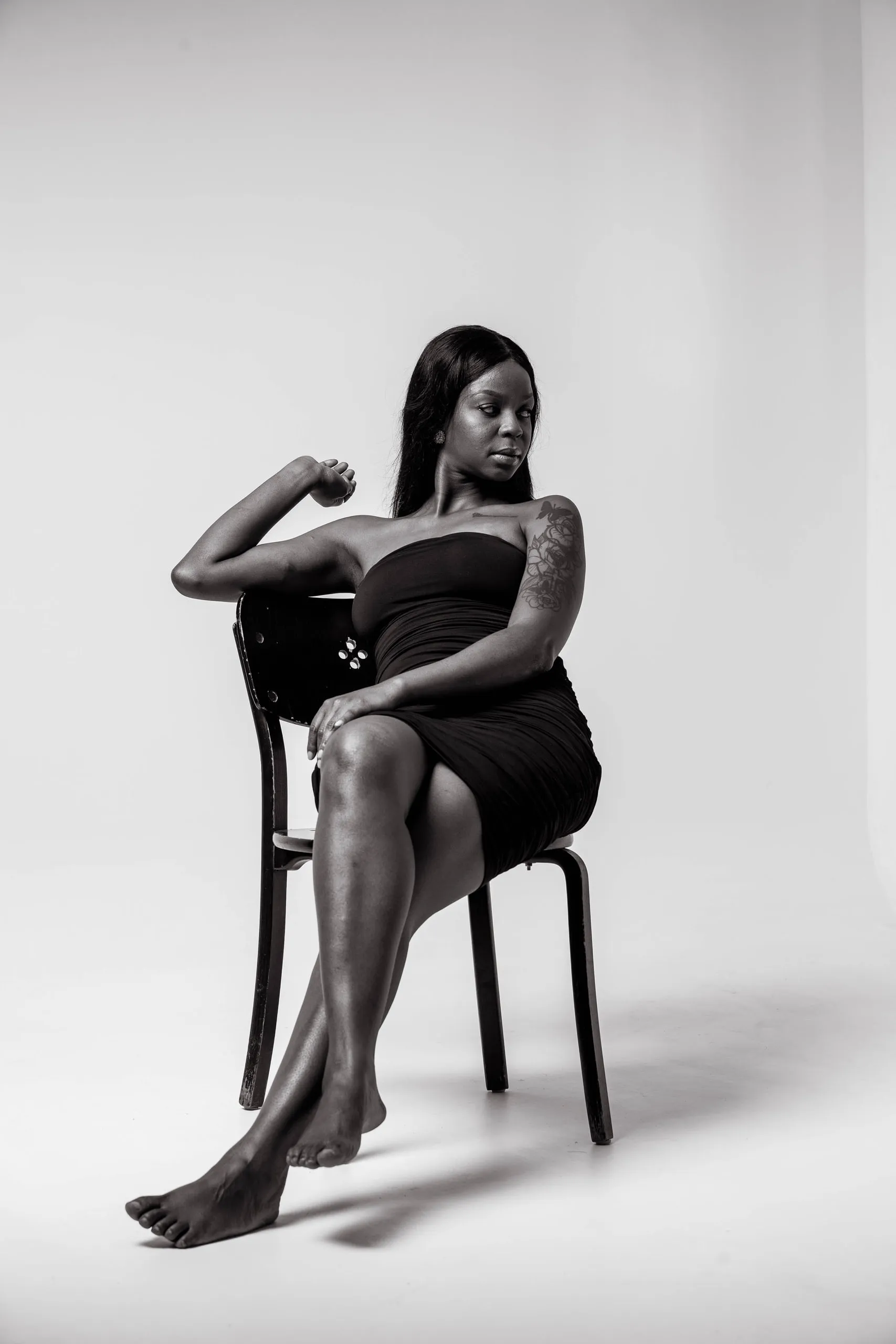 Woman in a black strapless dress sitting on a chair with one arm raised, in black and white