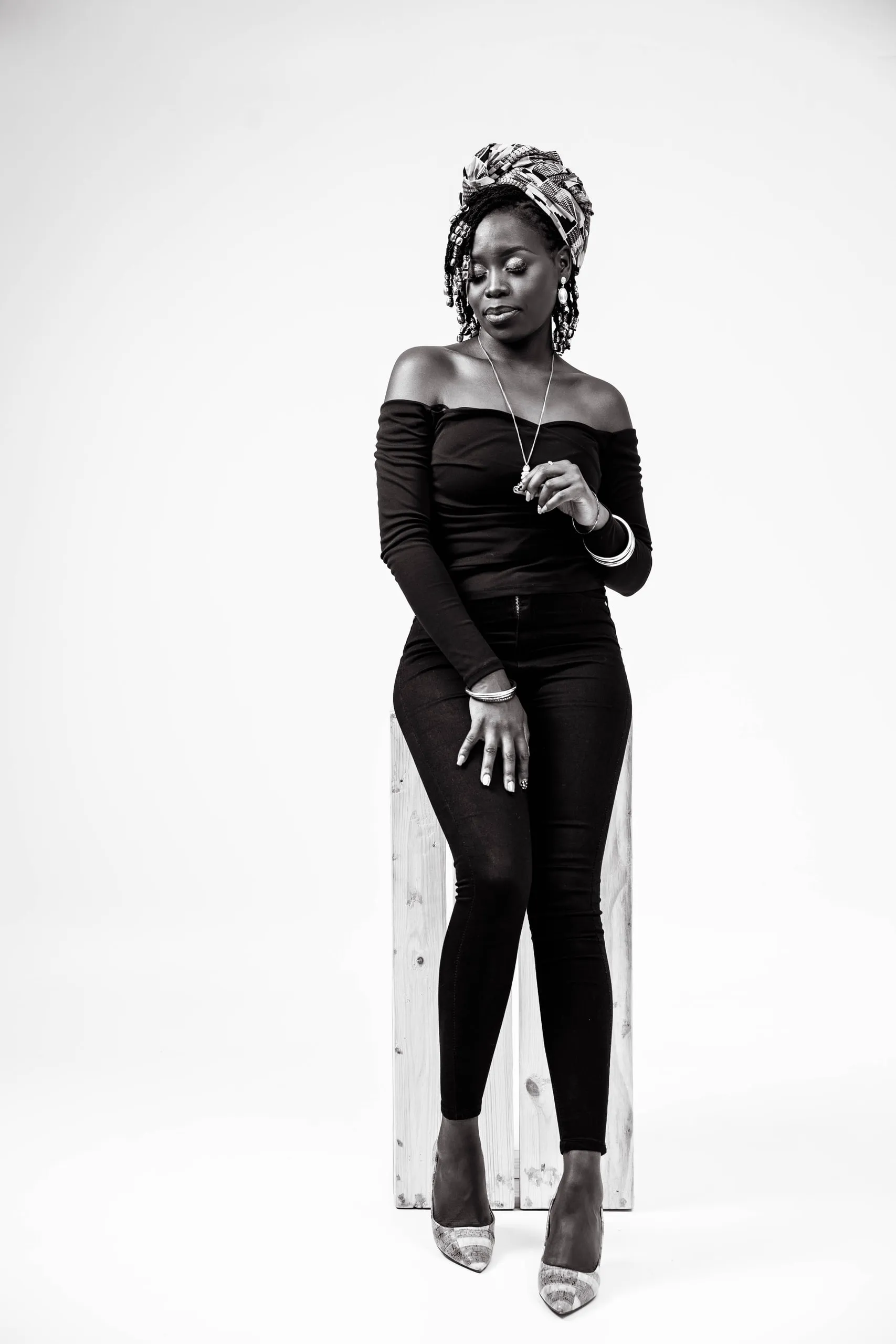 Woman in a black off-shoulder top and head wrap standing by a wooden crate, in black and white