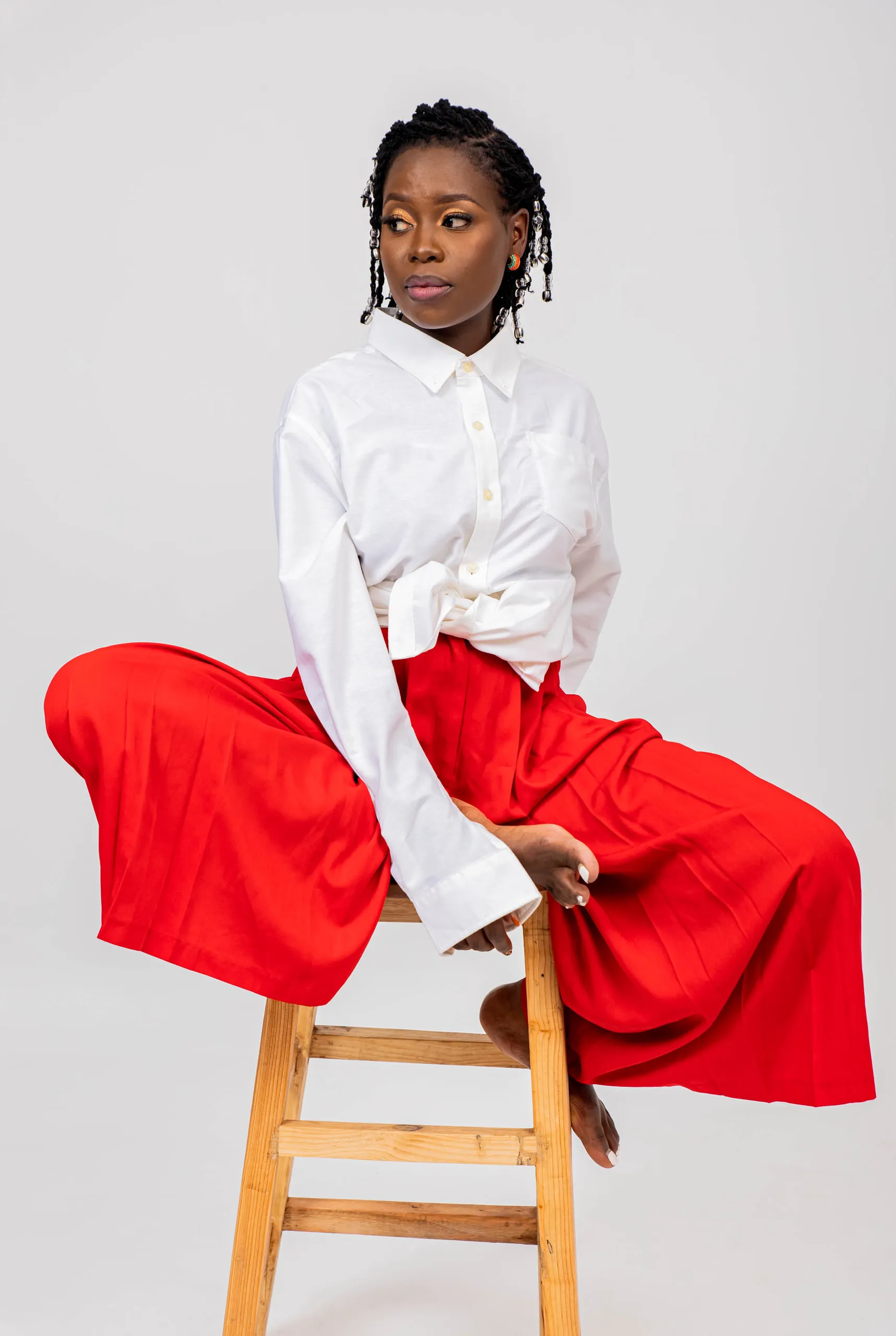 Woman in a white shirt and wide red trousers perched on a wooden stool, looking over her shoulder