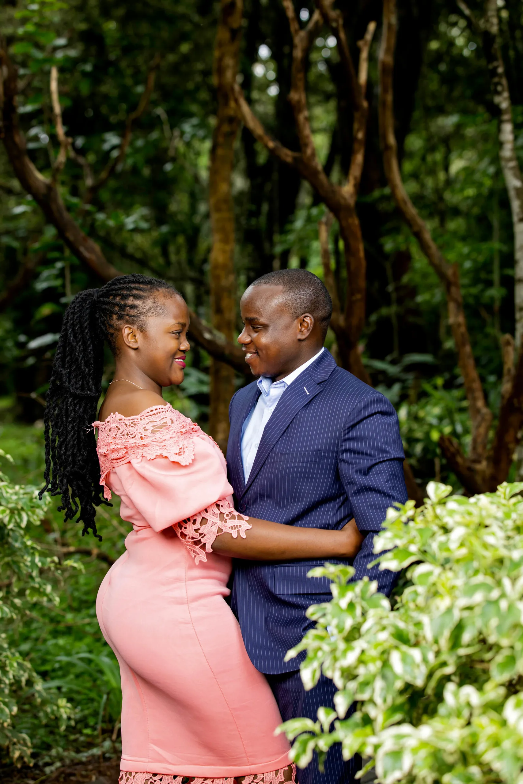 Couple smiling at each other in a forest, she in a pink lace dress
